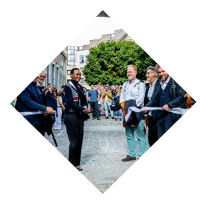 Een groep mensen staat buiten op een geplaveide straat met een grote boom op de achtergrond terwijl enkele personen een lint vasthouden, mogelijk voor een ceremonie of evenement.

Beschrijving gegenereerd door AI