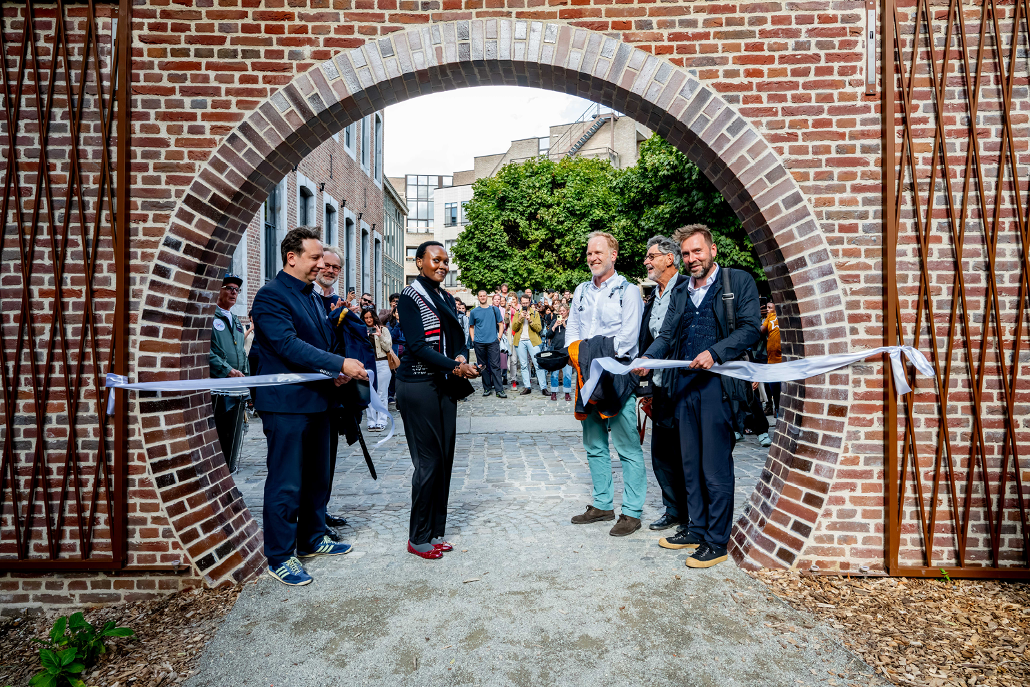 Een groep volwassenen staat bij een ronde bakstenen poort waar een lint wordt doorgeknipt tijdens een feestelijke opening in een stedelijke omgeving.

Beschrijving gegenereerd door AI